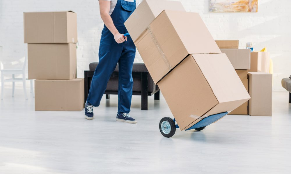cropped view of mover in uniform transporting cardboard boxes on hand truck in apartment